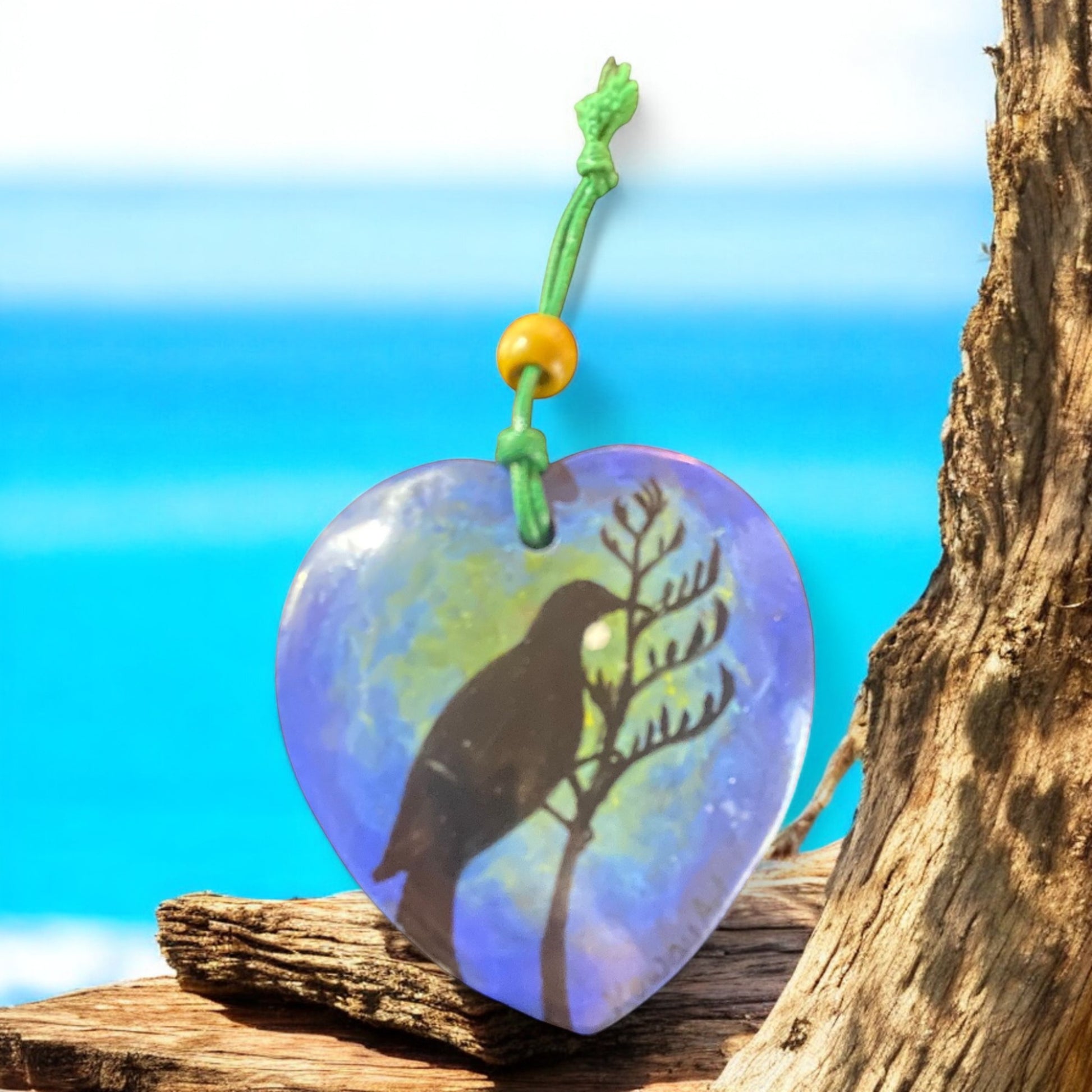 Heart-shaped pendant with Tui bird and branch design on a wooden surface with a beach background