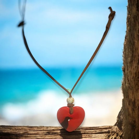 Red heart-shaped pendant showing Tui Bird on a brown cord against a beach background