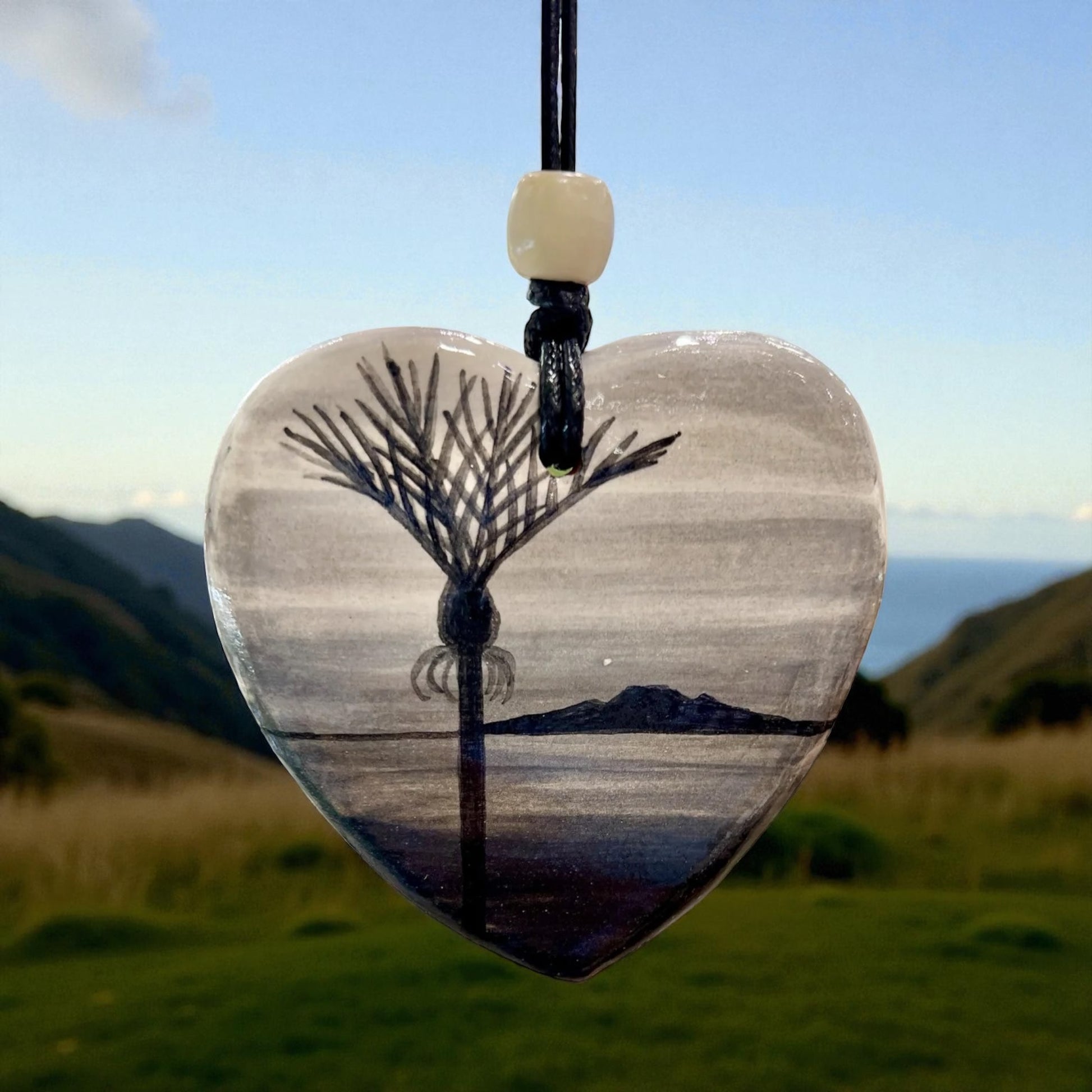 Heart-shaped pendant with aNikau Palm reflection on water against a mountain backdrop